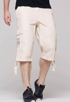 Beige Cargo-Shorts mit Taschen, Kordelzugsäumen und Stickdetails. Kombiniert mit schwarzen Sportschuhen. Der Stoff wirkt robust und strukturiert.