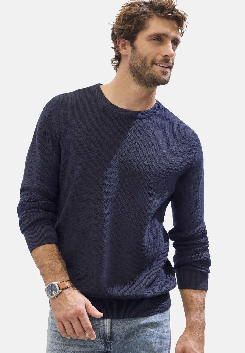 Marineblauer Strickpullover mit rundem Halsausschnitt, gerippten Bündchen und Saum. Getragen mit hellblauen Jeans und einer braunen Lederarmbanduhr.