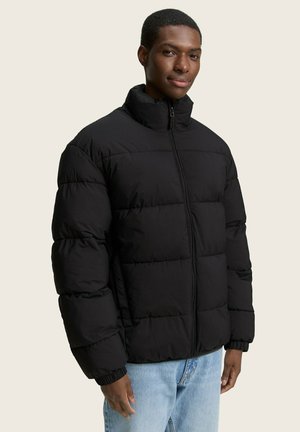 Chaqueta acolchada negra con cuello alto, cierre de cremallera y puños elásticos. Textura suave con un acabado liso, presentando una silueta cuadrada.
