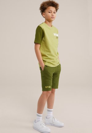 Grön bomullströja med korta ärmar, med en mörkare grön detalj. Matcher med matchande shorts och vita sneakers.