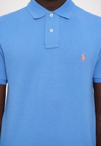 Gros plan d'une personne portant un polo bleu clair avec deux boutons blancs et un logo brodé orange représentant un cheval sur la poitrine gauche.