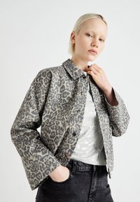 Veste en imprimé léopard dans des tons gris, avec un col, une fermeture à boutons, et des manches larges, associée à une chemise argentée à motifs et un jean foncé.