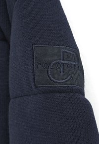 Suéter de punto azul marino con un acabado texturizado y un parche del logo bordado en negro en la manga. Primer plano del tejido y los detalles del diseño.