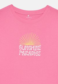Camiseta de algodón rosa con cuello redondo, mangas cortas y un diseño gráfico que presenta "SUNSHINE PARADISE" en blanco con un patrón de estallido amarillo.