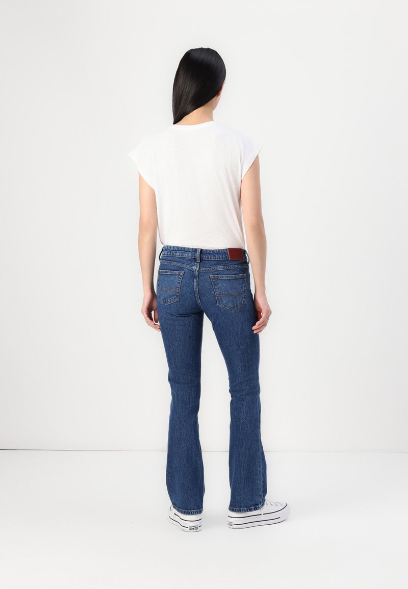 Weißes T-Shirt mit kurzen Ärmeln kombiniert mit dunkelblauen Schlagjeans, die zwei Gesäßtaschen und einen Lederpatch aufweisen. Das Model trägt weiße Sneaker.