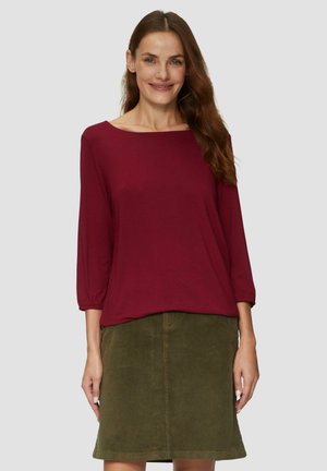 Bordeaux vrijzittend shirt met driekwart mouwen, gecombineerd met een olijfgroene corduroy rok. Heeft een ontspannen halslijn en een soepele textuur.