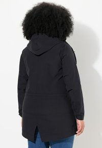 Svart vattentät jacka med huva, samlad midja och detalj med bakslits. Har ett glatt tyg och långa ärmar med elastiska manschetter.