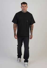 Czarny T-shirt z krótkim rękawem o luźnym kroju i małym logo na piersi, zestawiony z ciemnymi dżinsami i białymi sneakersami. Gładka tekstura materiału.