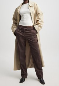 Beiger Trenchcoat über einem weißen, figurbetonten Oberteil, kombiniert mit schokoladenbraunen, maßgeschneiderten Hosen und schwarzen, spitzen Schuhen. Glattes Material, strukturierte Passform.