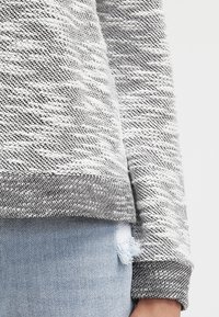 Grau-weiß gemusterter Pullover mit geripptem Saum und Bündchen, mit einem rohen Saumdetail an der Unterseite, kombiniert mit lässiger, abgenutzter Jeans.