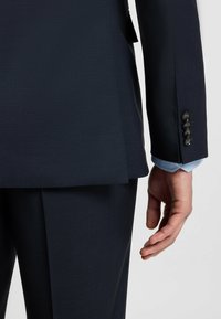 Blazer e pantaloni blu navy in tessuto testurizzato, con dettagli ai polsini a sei bottoni e un polsino di camicia azzurro chiaro che spunta sotto il blazer.