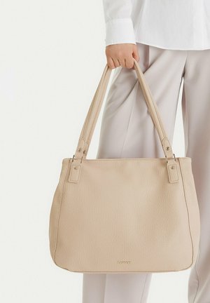 Person hält eine beige Lederhandtasche mit zwei Schulterriemen, trägt ein weißes Hemd und hellbeige Hose vor einem schlichten Hintergrund.