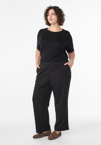 Melnas īsroku krekls kopā ar melnām platām biksēm, izgatavots no gludas auduma ar sānu kabatām; modelis valkā brūnas zamšādas mokasīnus.