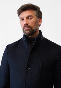 Marinblå ullblandad kappa med hög krage, tvåknappsstängning och en framficka. Har en mjuk textur och kontrasterande axeldetaljer.
