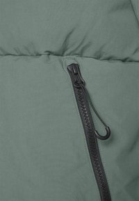 Grøn polstret overtøj med en sort lynlås og en trækflap. Materialet er glat og mat, med en quilted struktur synlig over lynlåsen.