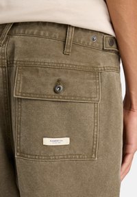 Olivgrüne Hosen mit einer Gesäßtasche, die über einen Druckknopfverschluss verfügt, sowie einem Etikett, das "ELEMENT CO. Since 1992." liest. Aus strukturiertem Stoff gefertigt.