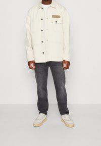 Chaqueta color crema con botones negros y un bolsillo que presenta una etiqueta beige; combinada con pantalones vaqueros oscuros y zapatillas blancas con suelas beige.