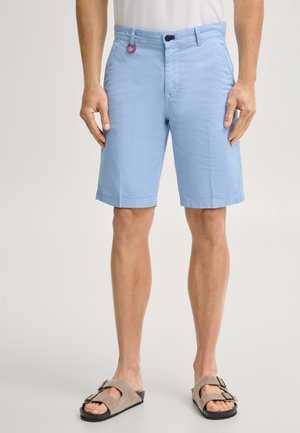 Man draagt lichtblauwe shorts met zijzakken en beige sandalen met dubbele bandjes, staand tegen een effen achtergrond.