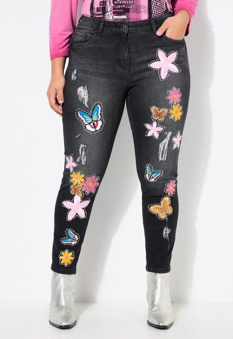 Jeans de mezclilla negros con coloridos bordados de mariposas y flores, que presentan detalles desgastados y un diseño ajustado. Botines plateados visibles.