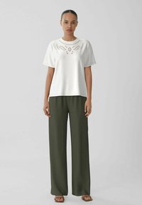 Top blanco de manga corta con diseño bordado en el cuello, combinado con pantalones holgados de color verde oliva. El modelo lleva sandalias marrones.