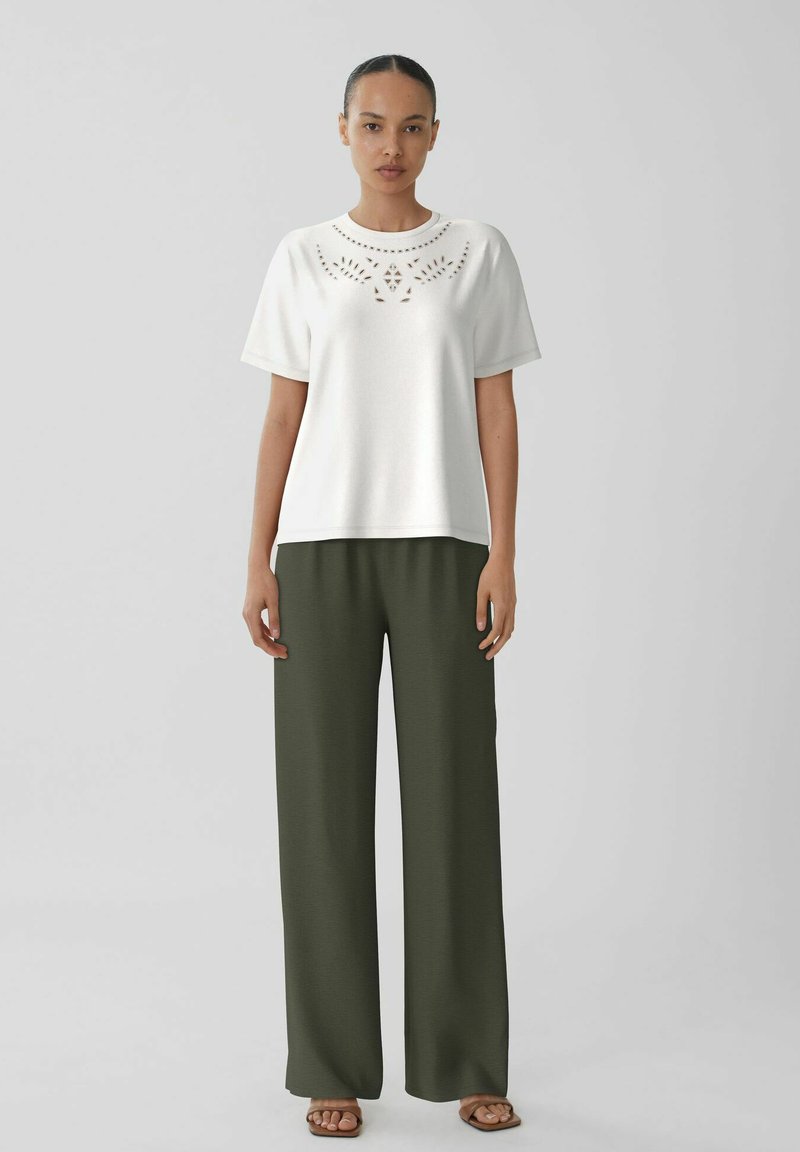 Top blanco de manga corta con diseño bordado en el cuello, combinado con pantalones holgados de color verde oliva. El modelo lleva sandalias marrones.