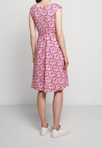 Růžové květinové šaty s krátkými rukávy, vypasovaným pasem a rozšířenou sukní. Tkanina má vzor listů v odstínech růžové a zelené. Bílé tenisky.