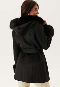 Cappotto nero con cappuccio e polsini foderati in pelliccia, vita aderente, e design sartoriale. Tessuto liscio con cuciture strutturate e vestibilità rilassata.
