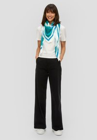 Turquoise en witte gemusterde sjaal over een wit T-shirt, gecombineerd met losse zwarte broek en witte sneakers. Soepele stof, casual pasvorm.