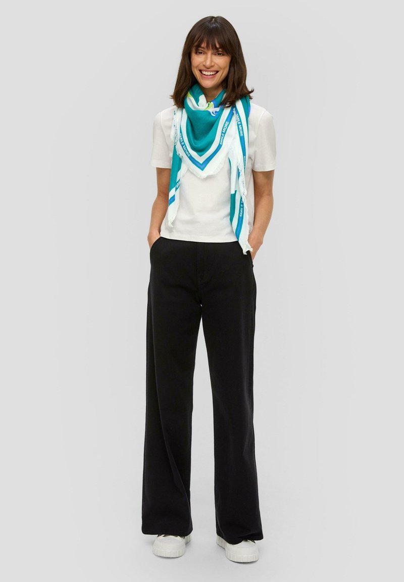 Turquoise en witte gemusterde sjaal over een wit T-shirt, gecombineerd met losse zwarte broek en witte sneakers. Soepele stof, casual pasvorm.