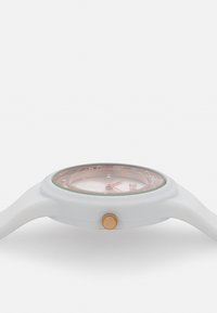 Reloj de silicona blanco con una caja redonda y transparente, un botón de acento dorado y una esfera rosa clara con marcadores y manecillas plateadas.