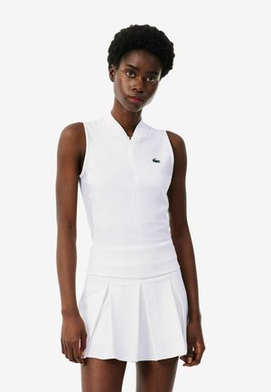 Vrouw draagt een witte mouwloze top met rits en een witte geplooide tennisrok, staand tegen een effen lichte achtergrond.