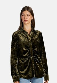 Camisa de terciopelo verde con mangas largas, cierre frontal de botones y detalle fruncido en la cintura. Presenta un cuello puntiagudo y una textura suave.