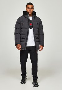 Zwarte pufferjack met capuchon, voorzien van een rits, dubbele voorzakken en rode accenten. Draagt over een graphic t-shirt en zwarte broek.