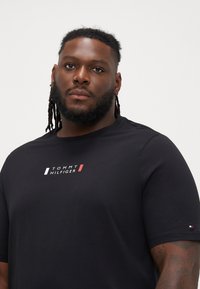 Μαύρη μπλούζα φτιαγμένη από βαμβάκι με στρογγυλή λαιμόκοψη, που διαθέτει λευκό και κόκκινο λογότυπο "TOMMY HILFIGER" στο στήθος και ένα μικρό λογότυπο στο μανίκι.