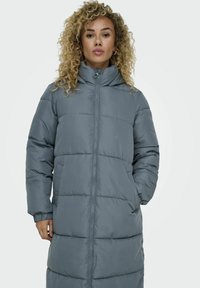 Manteau long gris rembourré avec un col haut, fermeture éclair à l'avant, poignets élastiques et poches latérales. Texture lisse et brillante avec des coutures horizontales.