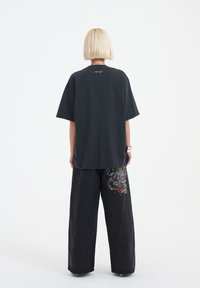 Schwarzes, übergroßes T-Shirt mit kurzen Ärmeln, das subtile Nähtdetails auf dem Rücken aufweist. Kombiniert mit weit geschnittenen, schwarzen Jeans, die mit bunten Drachenstickereien verziert sind.