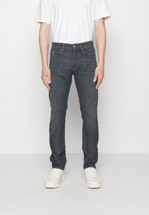 Męski model ubrany w ciemnoszare jeansy typu slim fit, białą koszulkę i białe trampki, stojący na jednolitym białym tle.