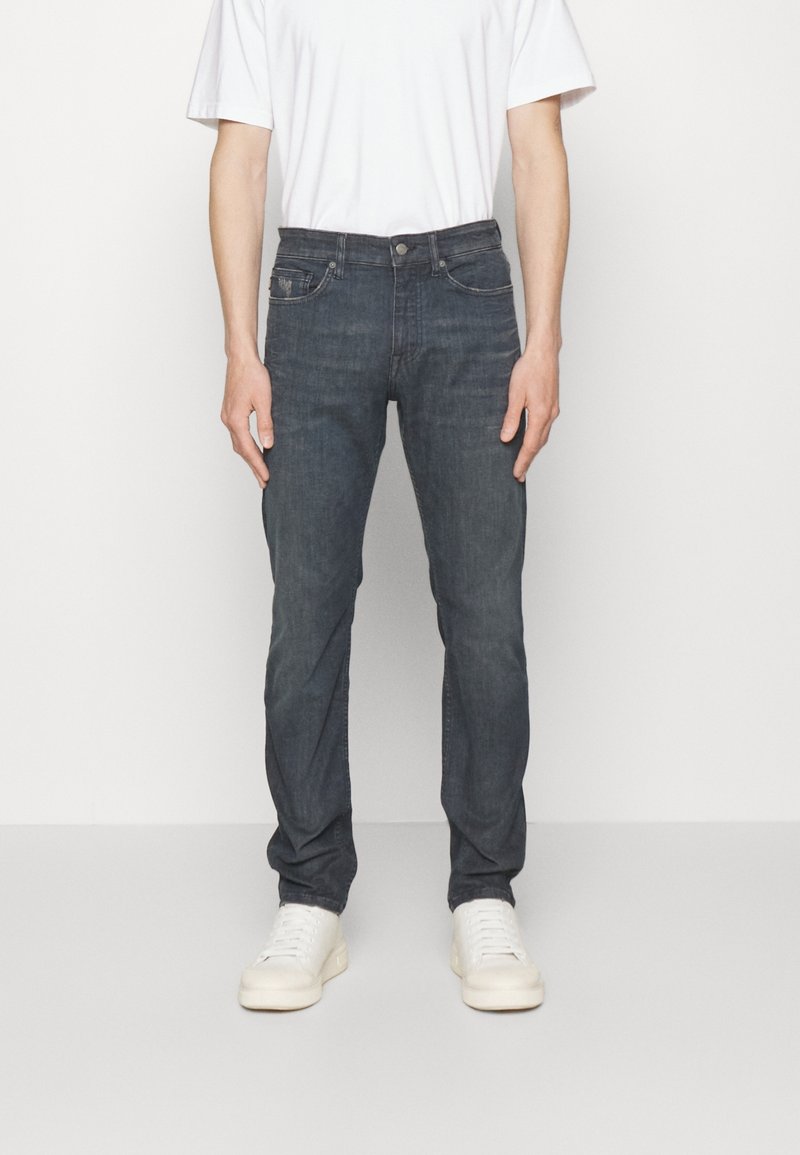 Modèle masculin portant un jean slim gris foncé, un t-shirt blanc et des baskets blanches, debout devant un fond blanc uni.