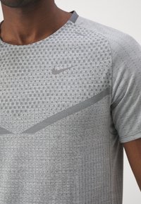 Šedé sportovní tričko Nike s texturovaným vzorem, kulatým výstřihem a krátkými rukávy. Obsahuje malé logo a akcenty geometrického designu.