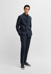 Granatowa dwurzędowa marynarka z klapami, w zestawieniu z pasującymi spodniami. Nosi ją z ciemną koszulą, uzupełniając całość ciemnymi sportowymi butami.