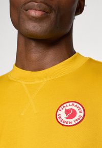 Sárga pulóver kerek piros folttal, amelyen egy róka logó és a "FJÄLLRÄVEN SWEDEN 1960" felirat található. Puha tapintású, kerek nyakkivágású és kötött mandzsettájú.