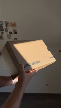 Ręce trzymają kartonowe pudełko po butach Salomon, a promienie słoneczne rzucają cienie w pokoju z rozproszonymi dekoracjami na ścianach.