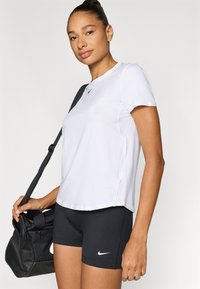 Λευκό αθλητικό πουκάμισο με κοντά μανίκια και στρογγυλή λαιμόκοψη με λογότυπο Nike, συνδυασμένο με μαύρο σορτς και μαύρη τσάντα τύπου duffel με λουράκι.