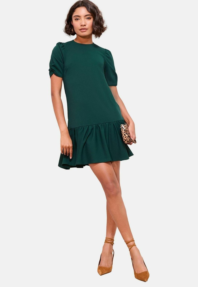 Rochie verde dintr-un material neted, cu mâneci scurte tip balon, tiv flared cu volane și un corset ajustat. Accesorizată cu o mică poșetă cu model.