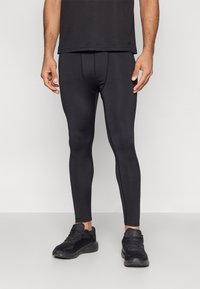 Žmogus vilki juodas aptemptas sportines tamprės, juodus bėgimo batelius ir juodą marškinėlį, stovi priešais neutralią šviesią fono sieną.