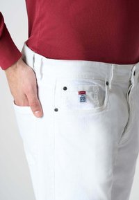 I pantaloni in denim bianco presentano un taglio classico con una tasca laterale e dettagli in metallo. La vita è a vista, abbinata a una maglietta bordeaux.