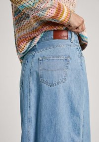 Svetlo modra denim krila z visokim pasom, ima dve zadnji žep in rjavo usnjeno oznako blagovne znamke na pasu, nad njo je teksturirana pletena jopica.