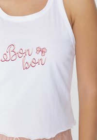 Bílý zkrácený top bez rukávů s textem "Bon bon" v pastelově růžovém kurzívě a malou růžovou grafikou mašličky. Měkká bavlněná tkanina s hladkou strukturou.