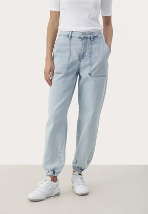 Lichtblauwe denim jeans met een relaxte pasvorm, voorzien van twee grote voorzakken en elastische boorden bij de enkels, gedragen met witte sneakers.