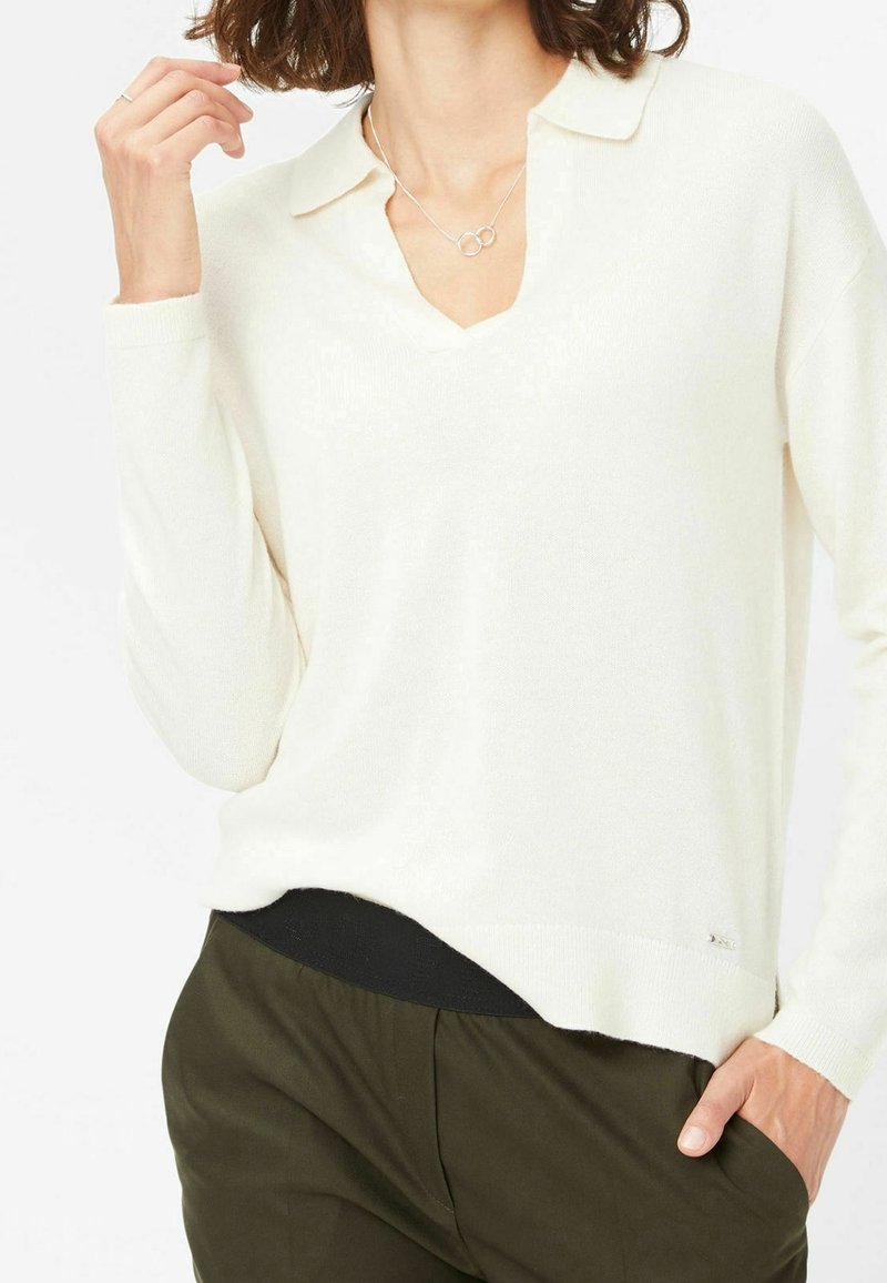 Femme portant un pull blanc à manches longues avec col, un pantalon vert olive et un collier en argent avec deux anneaux entrelacés.